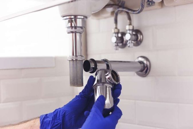 Un technicien en gants bleus répare le siphon sous un lavabo, en arrière-plan on aperçoit une vanne d'arrêt murale.