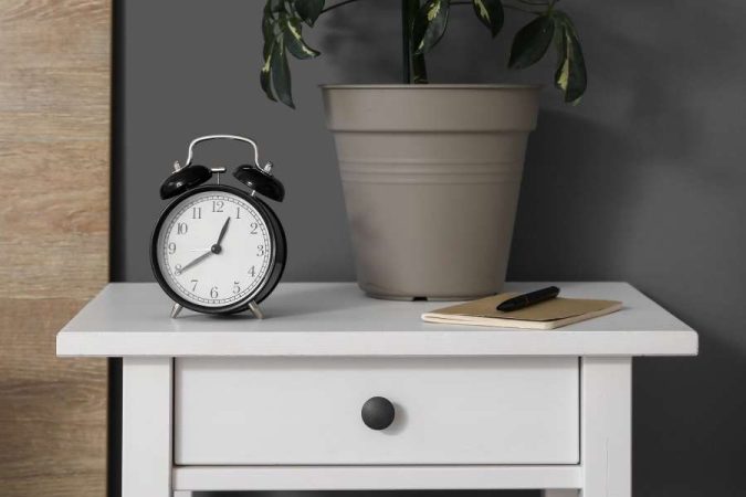 Une table de chevet suspendue blanche surmontée d'un réveil noir et d'un pot de fleurs.