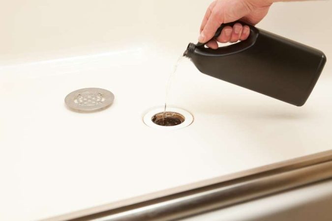 Une main verse un liquide de débouchage depuis un bidon noir dans l'orifice ouvert de l'évacuation d'une douche à l'italienne blanche, dont la grille est retirée.