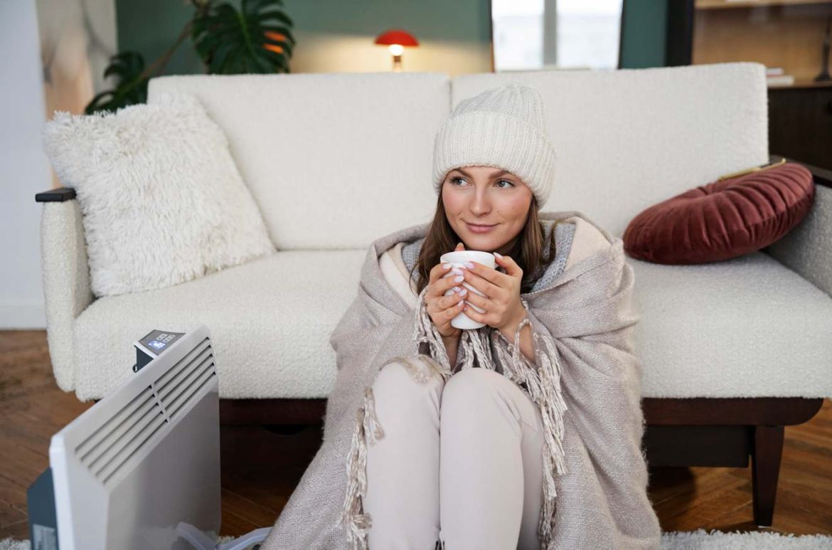 Une jeune femme emmitouflée dans un plaid et un bonnet tient une tasse chaude près d'un radiateur dans son salon, illustrant quel chauffage d appoint choisir pour passer l'hiver au chaud.